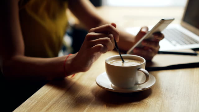 Close-up of woman's hands mixing coffee in cup and using smartphone relaxing in cafe