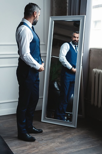 Elegant look. Full length of handsome man in full suit adjusting his jacket while standing in front of the mirror indoors