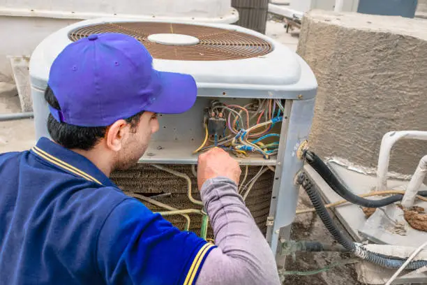 Photo of a professional electrician man is giving up thumbs up after the fixing a heavy duty air conditioner on the roof top and wearing blue uniform and cap and the city is in the background