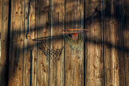 an old shabby basketball basket is throwing an evening shadow on a weathered wooden wall