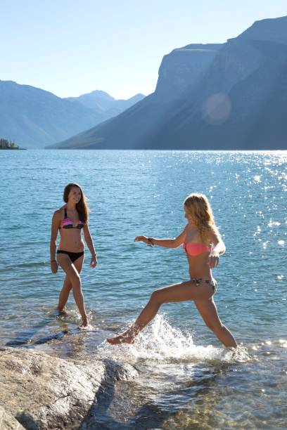 Two young women splash along a mountain lake shore They are in bikinis and they are playing in the water below the mountains splash mountain stock pictures, royalty-free photos & images