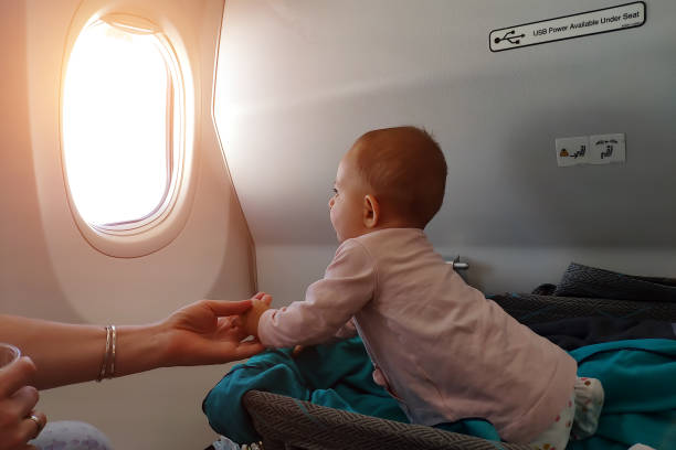 Happy infant baby lyes in special bassinet in airplane at his stomach. First flight of the baby, she is impressed and looking in the window of plane stock photo