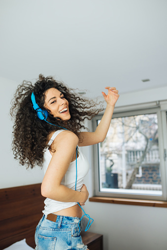 Happy Young Woman In Earphones Is Listening To Music With Smart Phone, Dancing And Smiling, Standing On The Bed