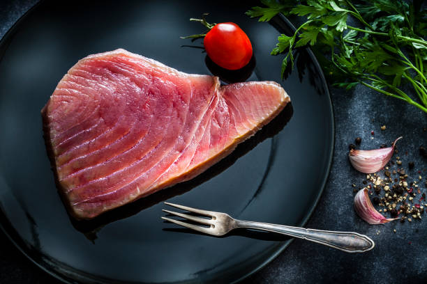 Raw food: top view of a fresh raw tuna steak on black plate shot on dark table. Some ingredients for cooking tuna steak like garlic, parsley, salt and pepper are all around the plate with the fillet. A fork is also included in the composition. Predominant colors are red and black. Low key DSRL studio photo taken with Canon EOS 5D Mk II and Canon EF 100mm f/2.8L Macro IS USM.