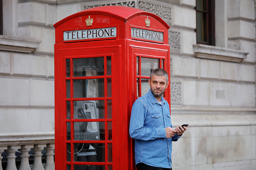 Young man using her smartphone, standing in front a phone booth
