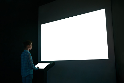Woman using electronic kiosk and looking at white blank large interactive wall display in dark room of modern technology exhibition. Mock up, futuristic, template, education and technology concept
