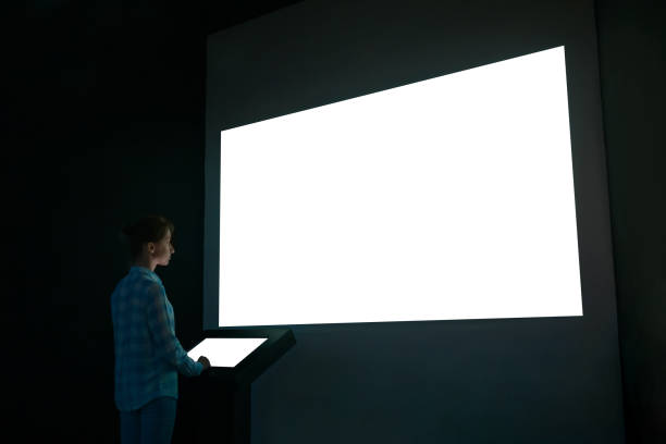 Woman looking at white blank large interactive wall display - mockup image Woman using electronic kiosk and looking at white blank large interactive wall display in dark room of modern technology exhibition. Mock up, futuristic, template, education and technology concept large screen stock pictures, royalty-free photos & images
