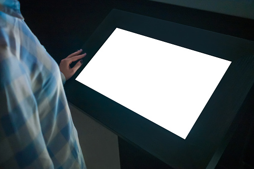 Woman looking at white blank interactive touchscreen display of electronic multimedia kiosk in dark room of technology exhibition - close up view. Mock up, copyspace, template and futuristic concept