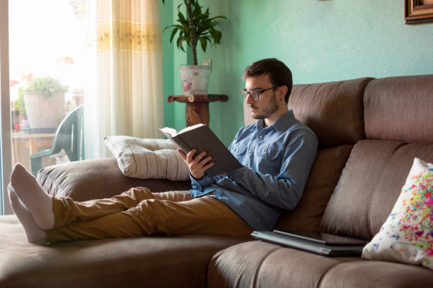 istock Young man with book on sofa at home 1160043641
