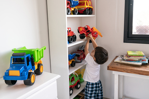 Cute little boy playing with his toy cars reaching one from shelf at home