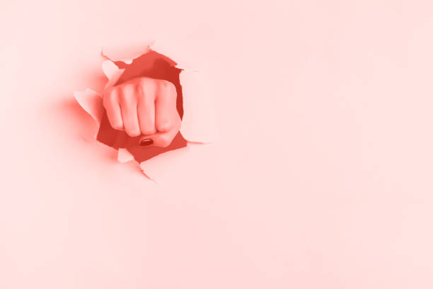 トレンディなサンゴのカラーペーパーの背景を通してパンチ女性の拳。戦争、闘争、紛争、フェミニストの概念。コピースペース付きバナー - strength struggle emotional stress business ストックフォトと画像