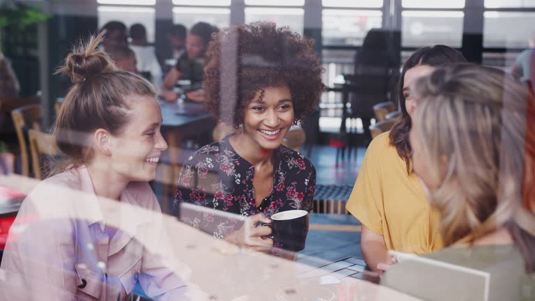 Group of young female friends meeting and sitting around table talking in cafe shot through window with reflections - shot in slow motion