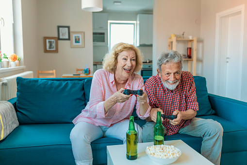 They have great game console.Happy senior couple playing video games. Senior couple having fun at home playing video game holding joysticks in hands sitting cozy on the sofa in bright sunny living room with big windows. Happy retirement concept.