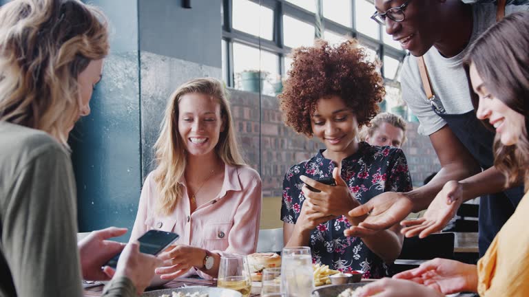 Group of young female friends meeting and sitting around table photos of food on mobile phones to post on social media as they are served by waiter - shot in slow motion