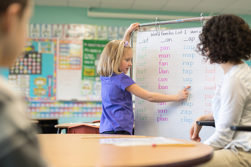 A first grader stands in front of a giant hanging sheet of spelling words and sounds them out in front of the class. The teacher, an female, and the other students are encouraging her.