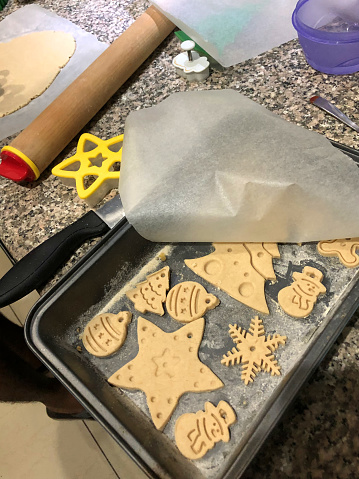 Stock photo of freshly baked Christmas cookies made in kitchen using cookie cutters, from salt dough to bake and paint by hand for Christmas tree decorations, oven tray tin with trees, gingerbread man / gingerbread men, stars, snowman / snow men