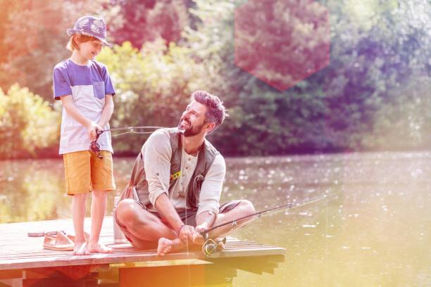 istock Smiling father looking at son standing on pier while fishing in lake 1160145963