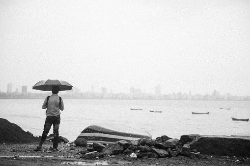 A man holding umbrella admiring distant skyline and enjoying rain near Marine Drive, Mumbai. Marine Drive is natural bay stretching 3.6 Km long, located in South Mumbai. When viewed from an elevated viewpoint or rooftop, the street lights resembles a string of pearls in a necklace and thus Marine Drive is often referred as the Queen's Necklace. Marine drive is loved among the locals and tourists.