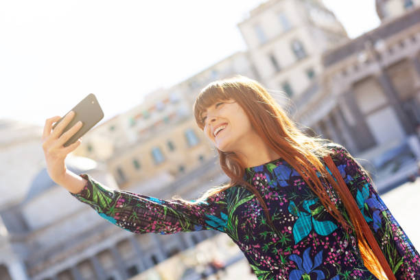 istock Smiling Young Woman Taking Selfie While Standing Against Building 1160166440
