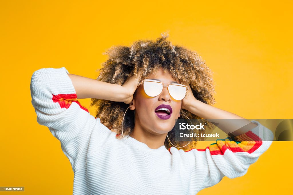 Funky afro girl against yellow background Portrait of funky young afro girl standing against yellow background with hand in hair and looking away Women Stock Photo
