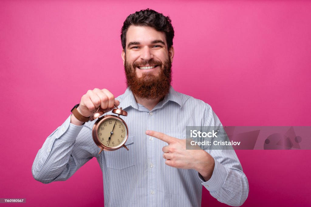 Smiling man with beard in casual pointing at alarm watch Clock Stock Photo