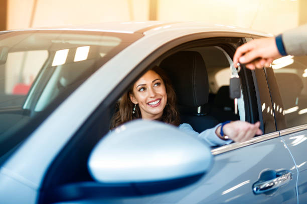 Car dealership. Buying new vehicle. Happy customer. An attractive brunette woman sitting in her brand new car and taking keys from vehicle dealer. car ownership photos stock pictures, royalty-free photos & images