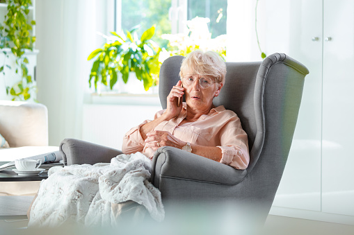Shot of an elderly woman sitting on arm chair looking worried while talking on mobile phone in her room