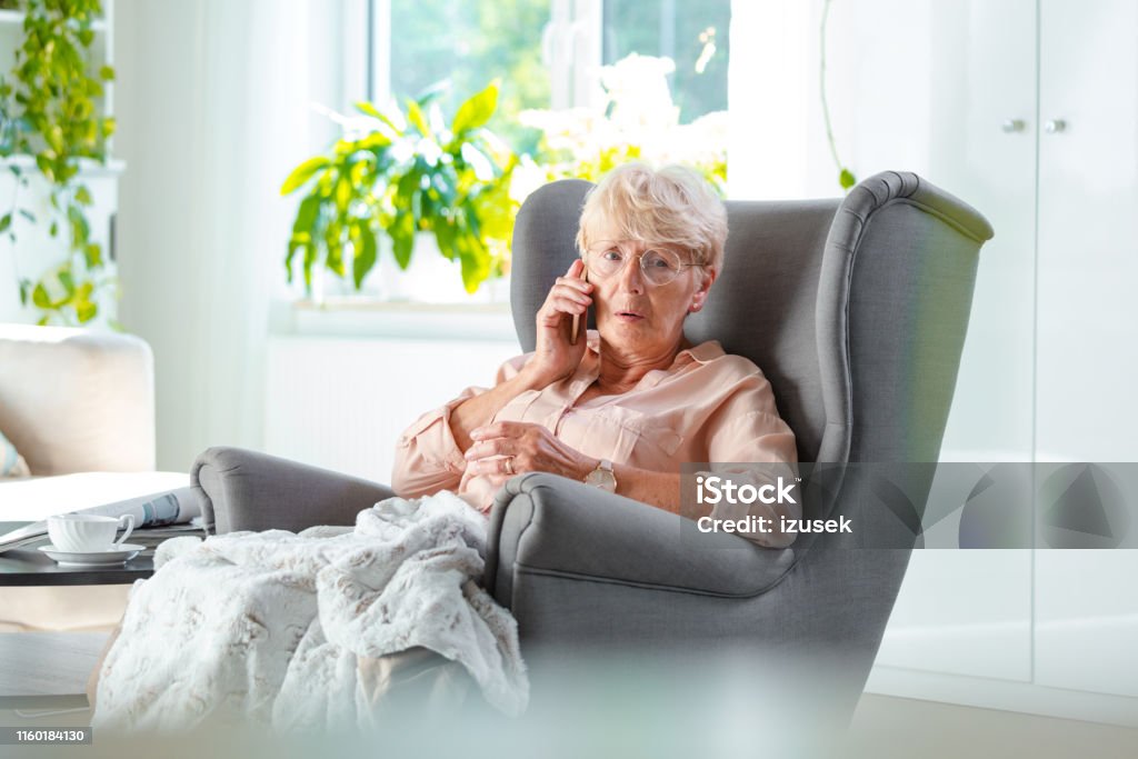 Elderly lady talking on cell phone looking worried Shot of an elderly woman sitting on arm chair looking worried while talking on mobile phone in her room Using Phone Stock Photo