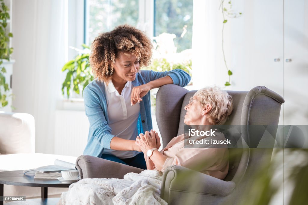 Caring nurse consoling an elderly lady Shot of young carer talking with senior woman sitting on arm chair in living room Senior Adult Stock Photo