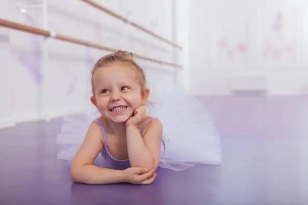 Adorable little ballerina girl laughing, looking away. Cute happy little girl in leotard and tutu skirt lying on the floor at ballet school, copy space