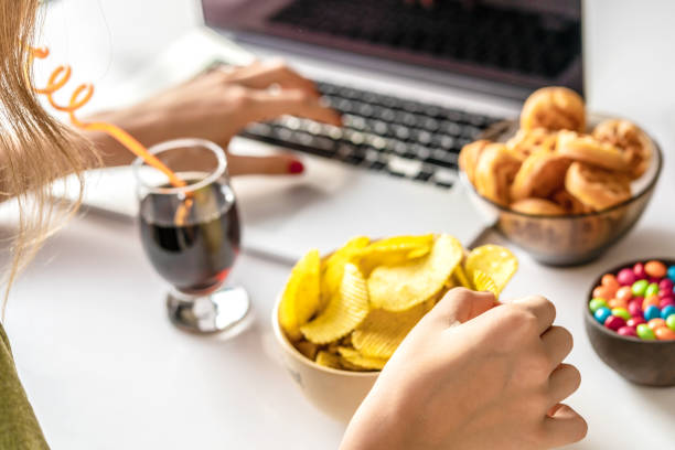 la ragazza lavora al computer e mangia fast food. cibo malsano: patatine, cracker, caramelle, cialde, cola. cibo spazzatura, concetto. - stuzzicare foto e immagini stock