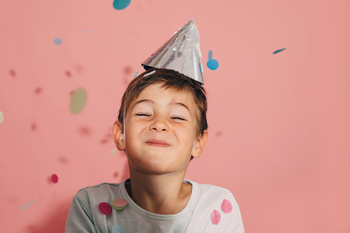 Photo of a cheerful boy with a celebration hat, celebrating his birthday while confetti's falling down. Studio shot.