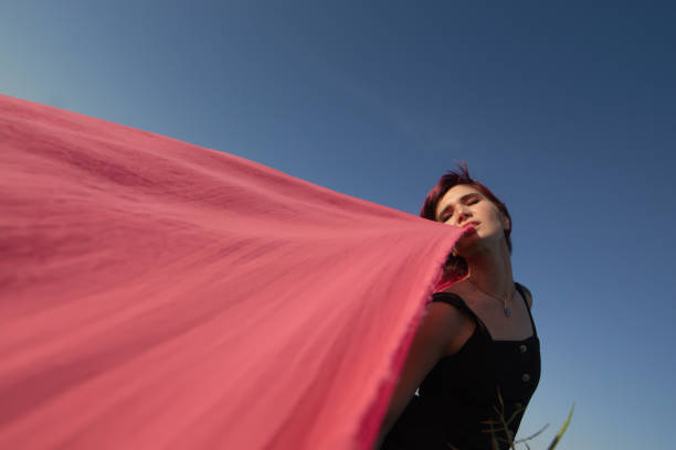 Cô gái trẻ tóc hồng với những mảnh vải màu hồng nhảy múa gần cánh đồng với máy phát điện gió. Cối xay gió lúc hoàng hôn. Chân dung thời trang