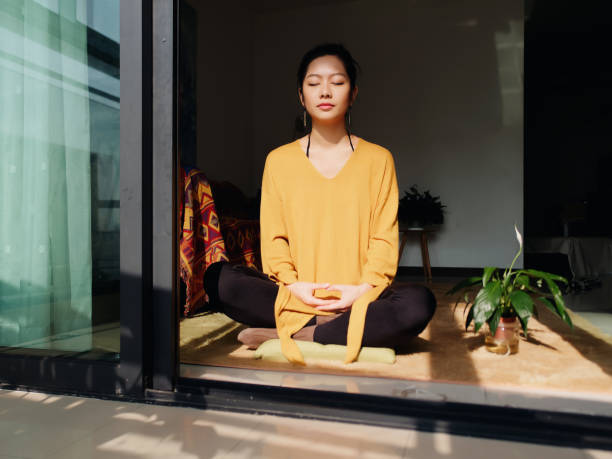 Pretty Chinese young woman meditating at home, sitting on floor with furry cushion in sun light, exercise, Lotus pose, prayer position, namaste, working out, Feeling peace and wellness concept. stock photo