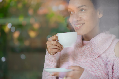 Gorgeous mixed race woman in pink turtleneck sweater standing in coffee shop, drinking espresso and looking trough window.