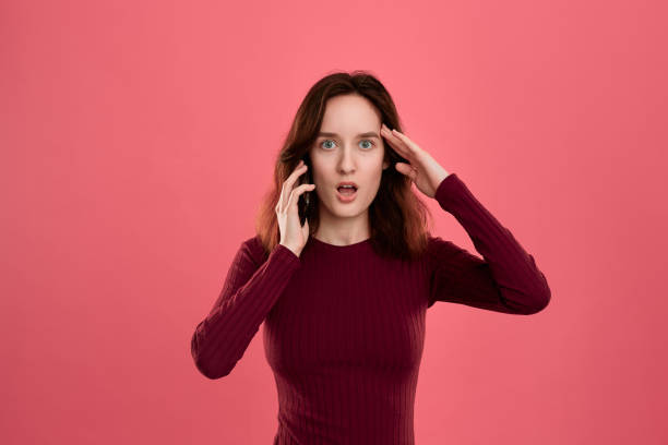 portrait of a beautiful brunette girl standing on a dark pink background and looking worried while speaking on the phone. - solicitous imagens e fotografias de stock