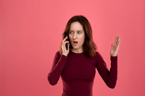 portrait of a beautiful brunette girl standing on a dark pink background and looking worried while speaking on the phone. - solicitous imagens e fotografias de stock