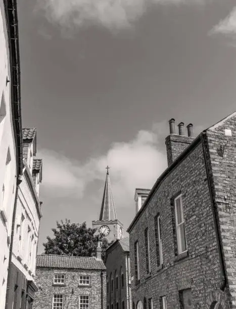 Wells Lane side street in Malton in Yorkshire. Old buildings are on each side and in the near distance.  A church spire, with a clock in the tower, is behind.