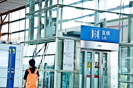 A traveling Chinese passenger walking past an elevator in the departure terminal of Beijing Capital International Airport (IATA: PEK, ICAO: ZBAA), Chaoyang–Shunyi, China. Mobile phone charging station nearby