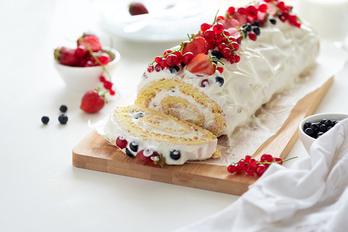 Sponge (biscuit) cake roll filling whipped cream and berries decorated strawberry, blueberry and red currants on white wooden background. Soft focus. Summer food concept