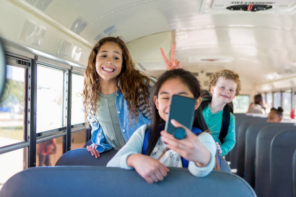 Playful friends on school bus Preteen schoolgirl takes selfie as her friend makes a rabbit ear gesture behind her. A young school boy is photobombing her selfie. photo bomb stock pictures, royalty-free photos & images