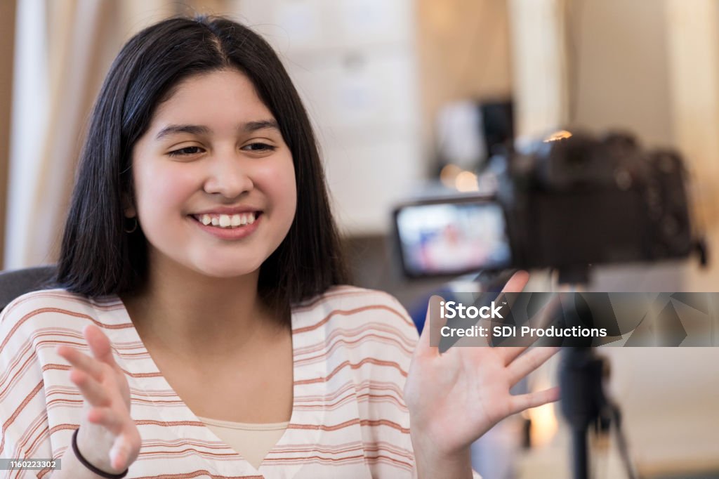 Teen girl smiles and gestures while filming with digital camera As she sits in the family workshop and uses the digital camera to do her homework, a teen girl smiles and gestures. Adolescence Stock Photo