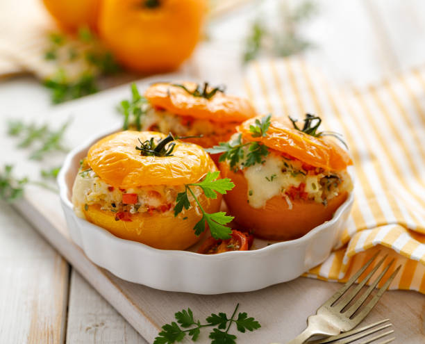 gefüllte tomaten, gebackene gelbe tomaten gefüllt mit bulgur, gemüse und käse mit dem zusatz von aromatischen kräutern in einer backform auf einem weißen holztisch. - stuffed tomato stock-fotos und bilder