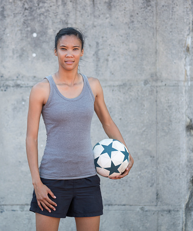Portrait of confident woman holding soccer ball. Young sports player is standing against wall. She is wearing gray tank top.