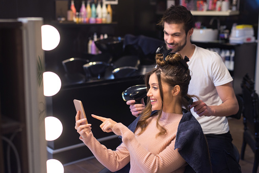 Young woman using smart phone while getting new hairstyle in the professional hair salon.