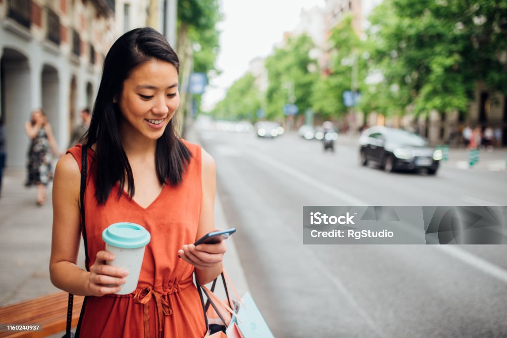 Young woman walking on the street Happy young woman walking on the city street after shopping and using her smartphone Using Phone Stock Photo