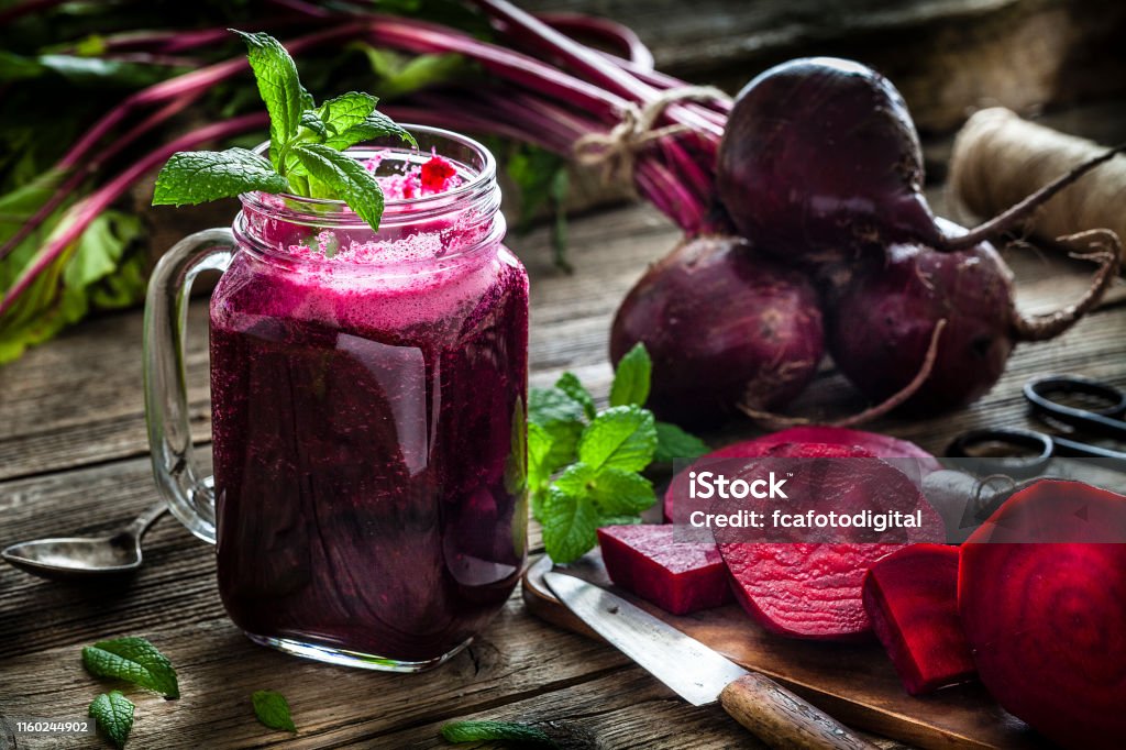 Healthy drink: beet juice on rustic wooden table Healthy drink: drinking glass filled with fresh organic beet juice shot on rustic wooden kitchen table. Whole and sliced beets are all around the glass. A kitchen knife, spoon and some mint twigs complete the composition. Predominant colors are purple, green and brown. Low key DSRL studio photo taken with Canon EOS 5D Mk II and Canon EF 100mm f/2.8L Macro IS USM. Common Beet Stock Photo