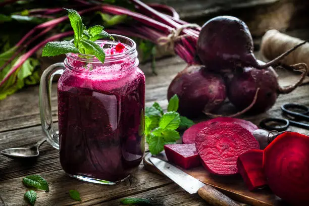 Healthy drink: drinking glass filled with fresh organic beet juice shot on rustic wooden kitchen table. Whole and sliced beets are all around the glass. A kitchen knife, spoon and some mint twigs complete the composition. Predominant colors are purple, green and brown. Low key DSRL studio photo taken with Canon EOS 5D Mk II and Canon EF 100mm f/2.8L Macro IS USM.