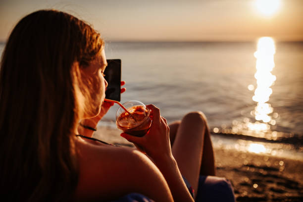 istock Teenage girl drinking coffee and using mobile phone on beach during beautiful sunset 1160261904