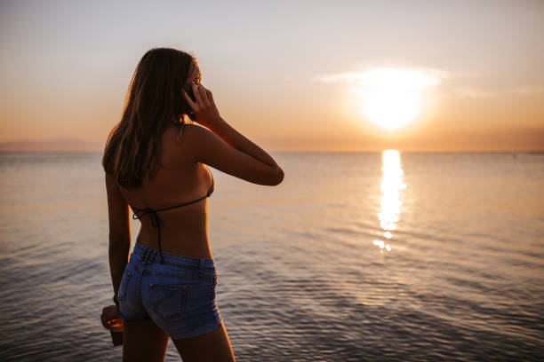 istock Young woman using smart phone on beach during idyllic sunset and holding nescafe 1160261910